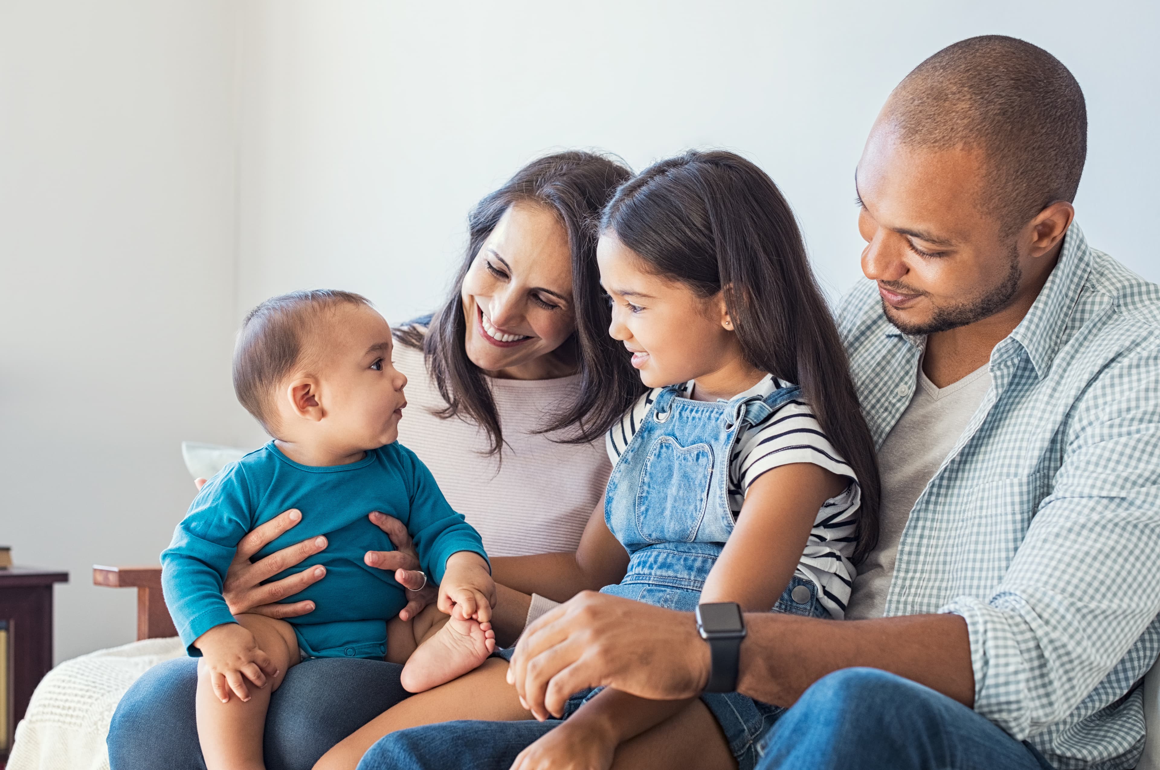 Multiethnic family playing with happy baby son at home. Parent and children relaxing together on the sofa at home in the living room. Little girl sitting on leg of dad looking her new cute brother.