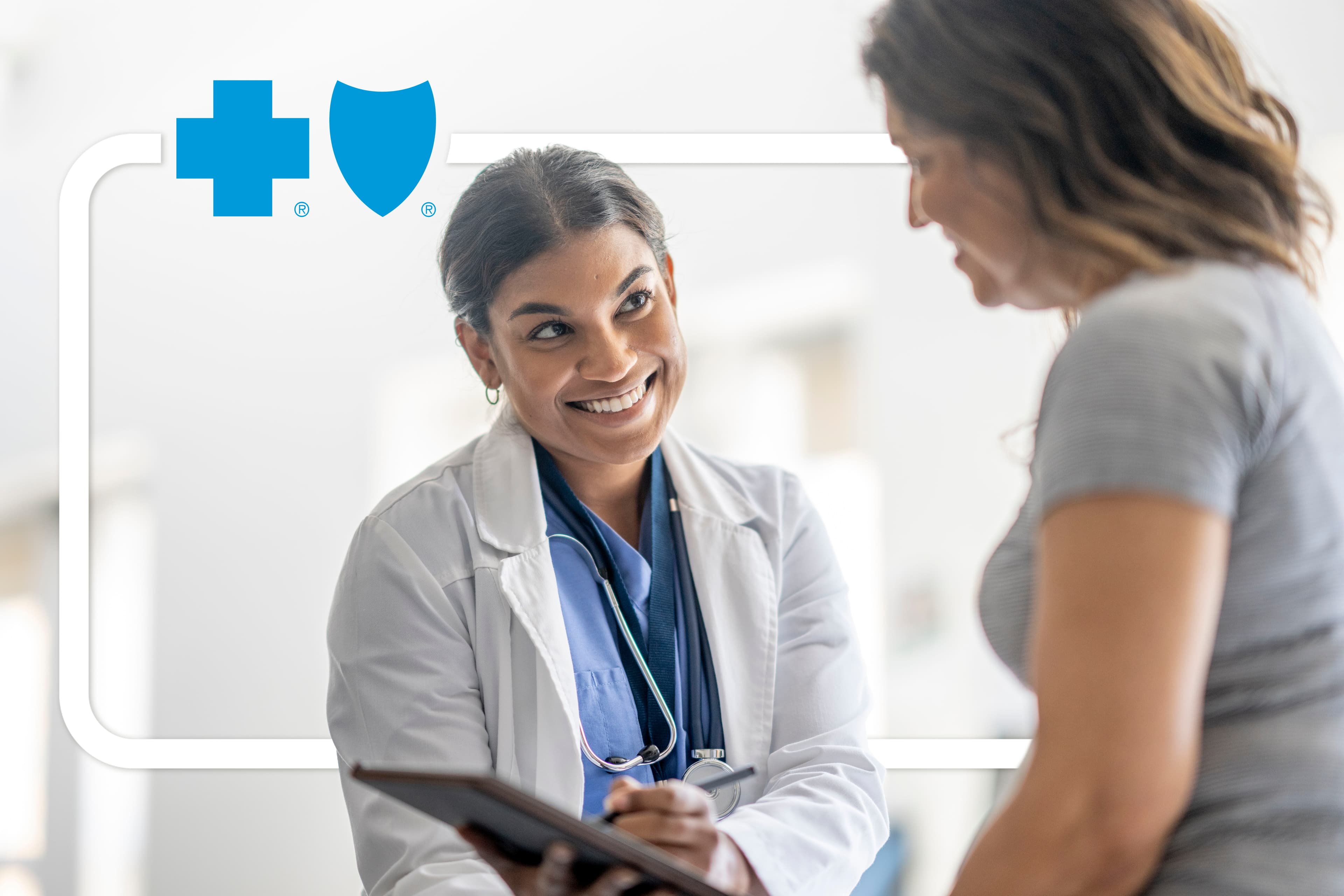 A female doctor sits across from a middle aged female patient as they meet to discuss her health.  She is dressed professionally in a white lab coat and has a clipboard in her hands as the two talk.