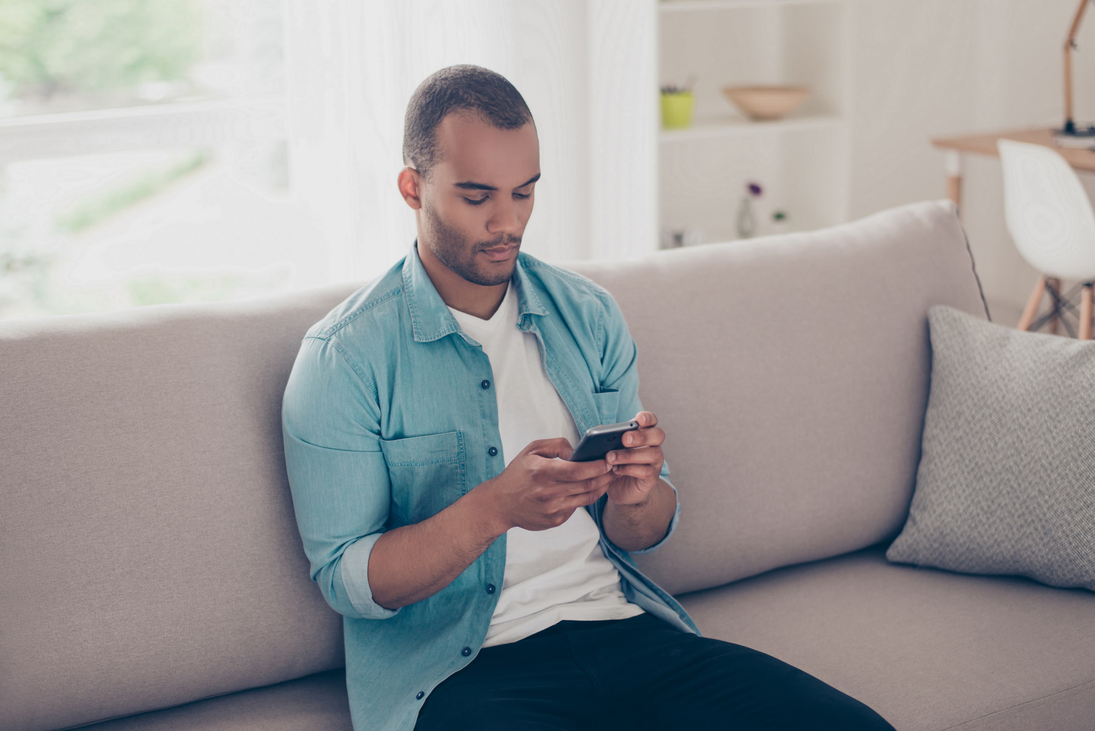 Close up shot of young concentrated black american guy, browsing on his smart phone, sitting on cozy beige sofa at home, in denim casual wear